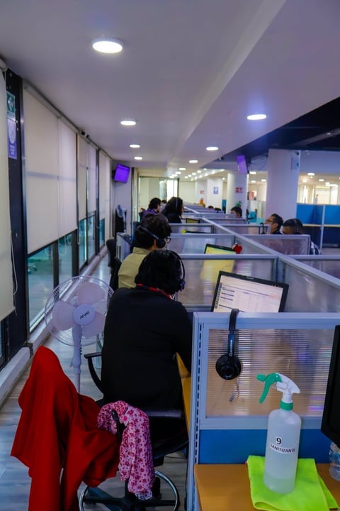 Office or call center workspace with multiple cubicles separated by translucent dividers, workers at desks with computers, hand sanitizer bottles visible, modern ceiling lights
