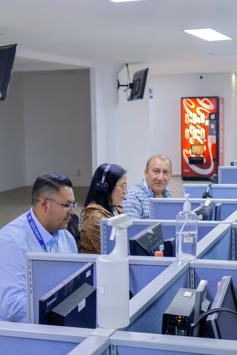 Three people sitting at a modern office desk in a bright room with white cubicles and an orange sign on the wall