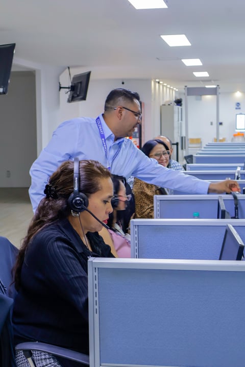 A supervisor instructs a female customer service agent wearing headphones at her cubicle in a busy call center office