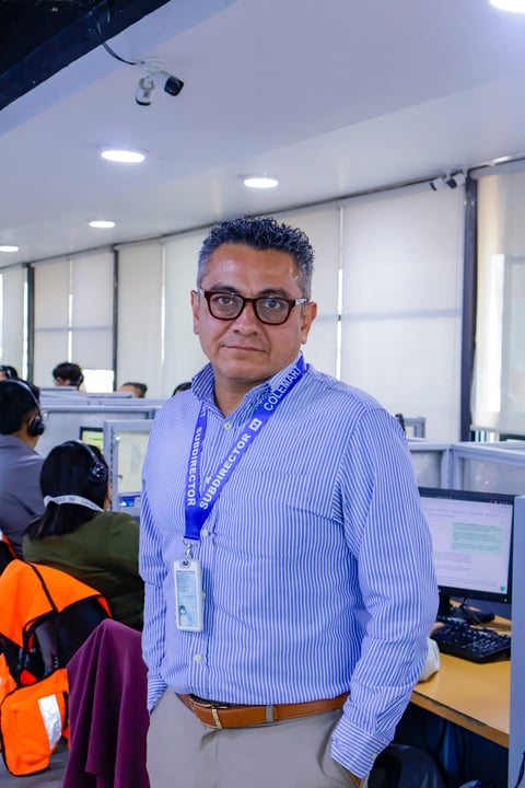 Man wearing glasses and blue striped shirt with blue ID badge standing in an office with colleagues in background