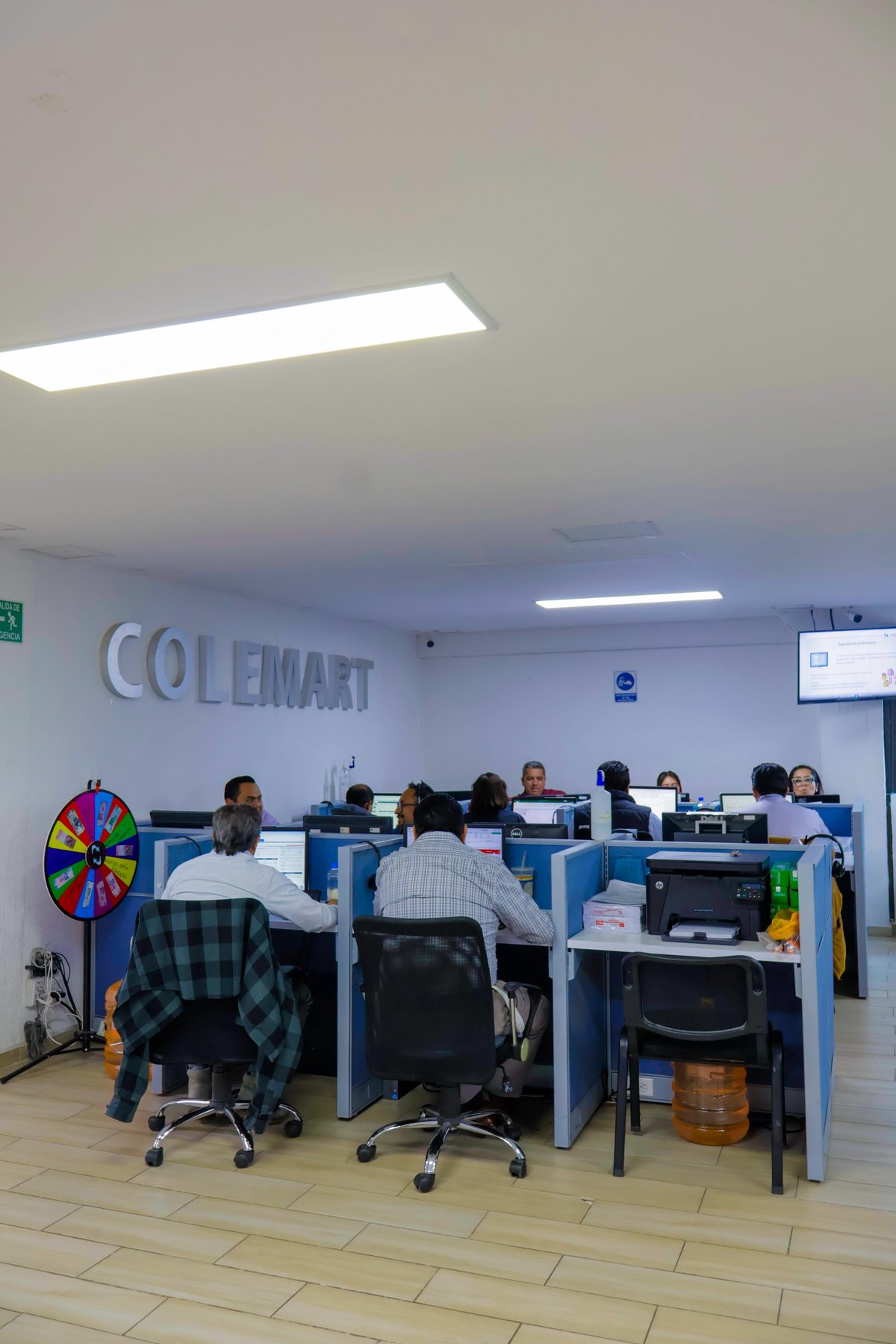Open office workspace with Colemart signage, multiple employees at blue desk cubicles with computers, modern lighting overhead