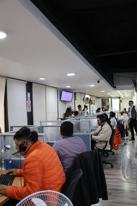 Interior of a busy office or service center with customers at desks and staff at a counter with a digital display screen
