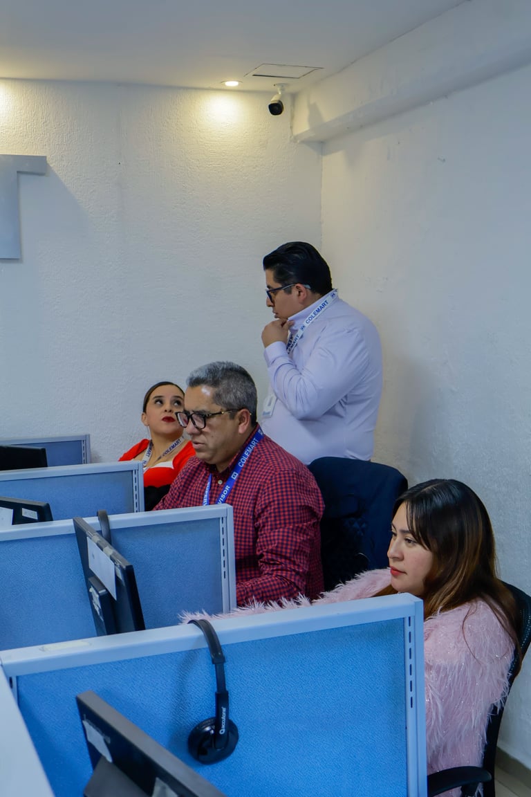 Four people in an office with blue desk partitions, reviewing work on computer monitors in a white room