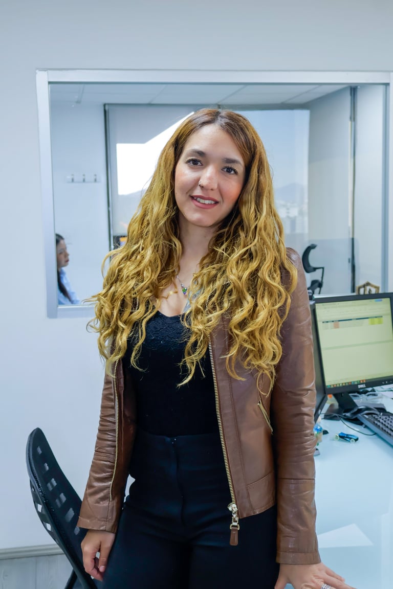 Young woman with long wavy blonde hair wearing a brown leather jacket over a dark top, standing in an office setting with a computer desk visible in the background