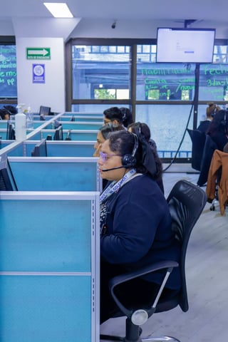 Call center employees wearing headsets working at blue cubicle desks in an office with windows and modern lighting