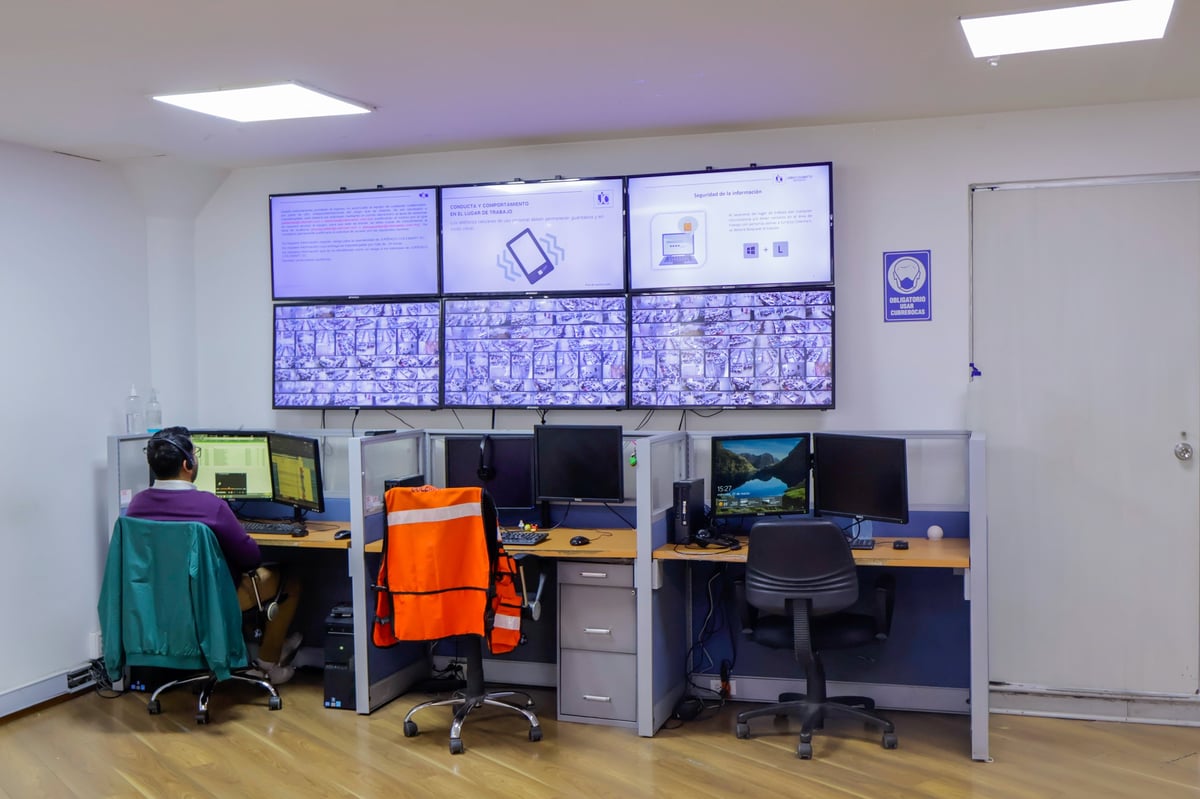 Modern office workspace with six computer monitors on wall above three organized cubicles with desks, chairs, and office equipment