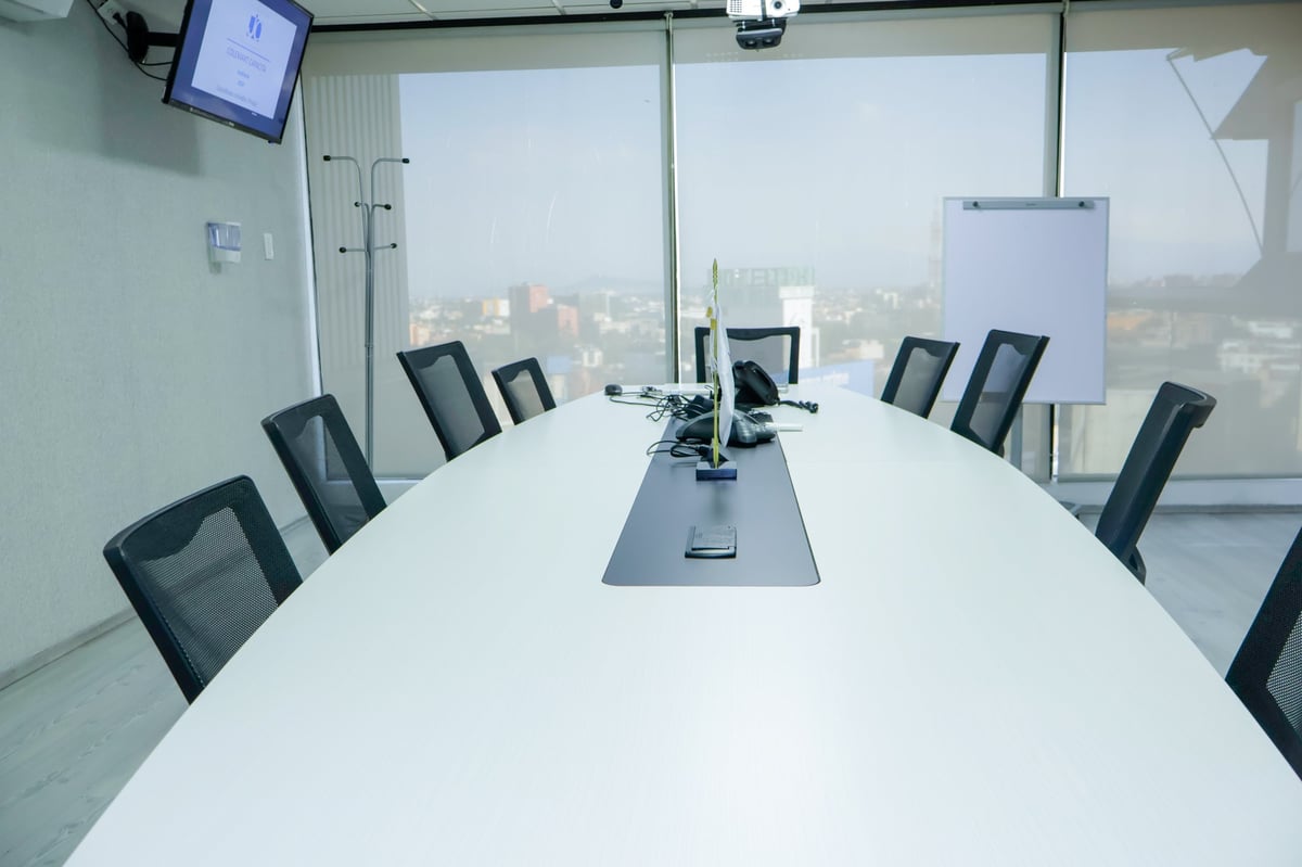 Sala de conferencias moderna con mesa blanca larga, sillas de malla negras y ventanales de piso a techo con vista al skyline de la ciudad