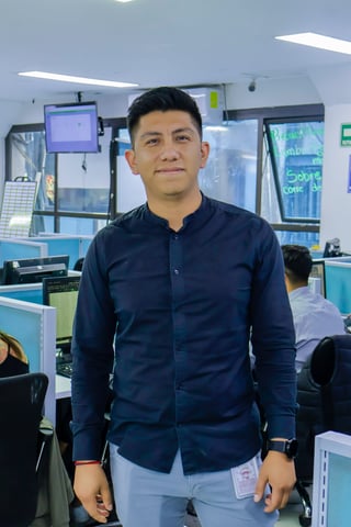 Man in dark blue button-up shirt standing in a modern office workspace with computer monitors and colleagues in the background