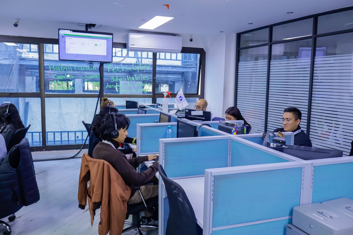 Modern office or service center with blue cubicles, workers at desks, customers waiting, and large windows overlooking an urban street