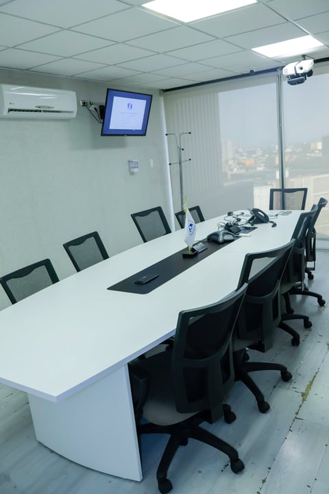 Modern office conference room with white curved table, black mesh chairs, wall-mounted screen, and floor-to-ceiling windows overlooking city