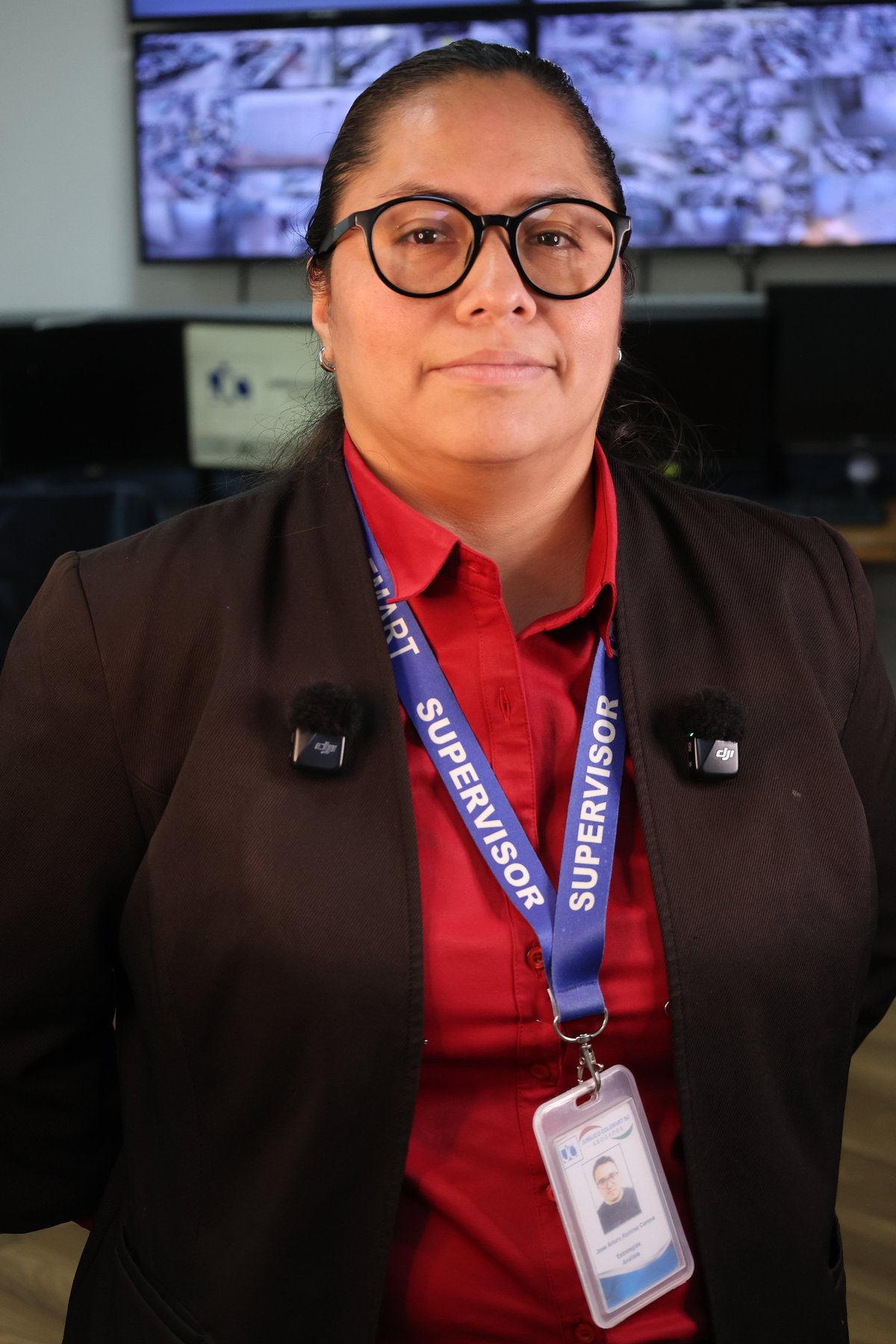 Professional woman wearing glasses, red shirt, brown blazer, and supervisor lanyard with ID badge standing in office.