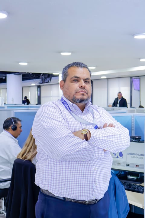 Man in light purple checkered shirt standing with arms crossed in modern office cubicle environment with colleagues in background