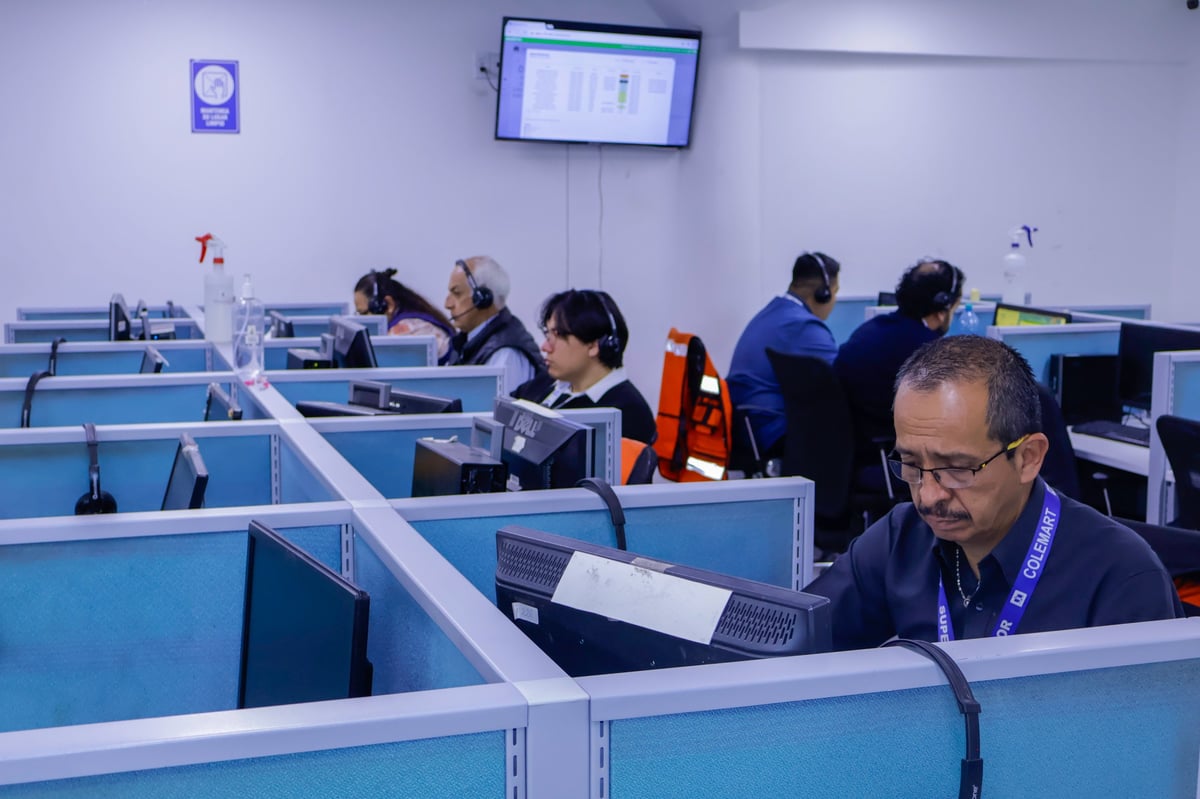 Workers in blue cubicles at a call center or office, with monitors and a clock visible on the wall.